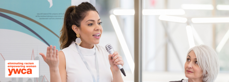 Woman speaking into microphone in front of crowd with another woman looking on