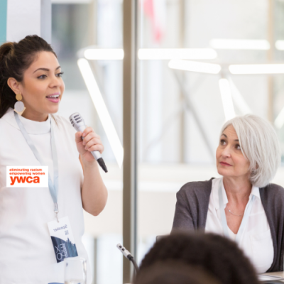 Woman speaking into microphone in front of crowd with another woman looking on